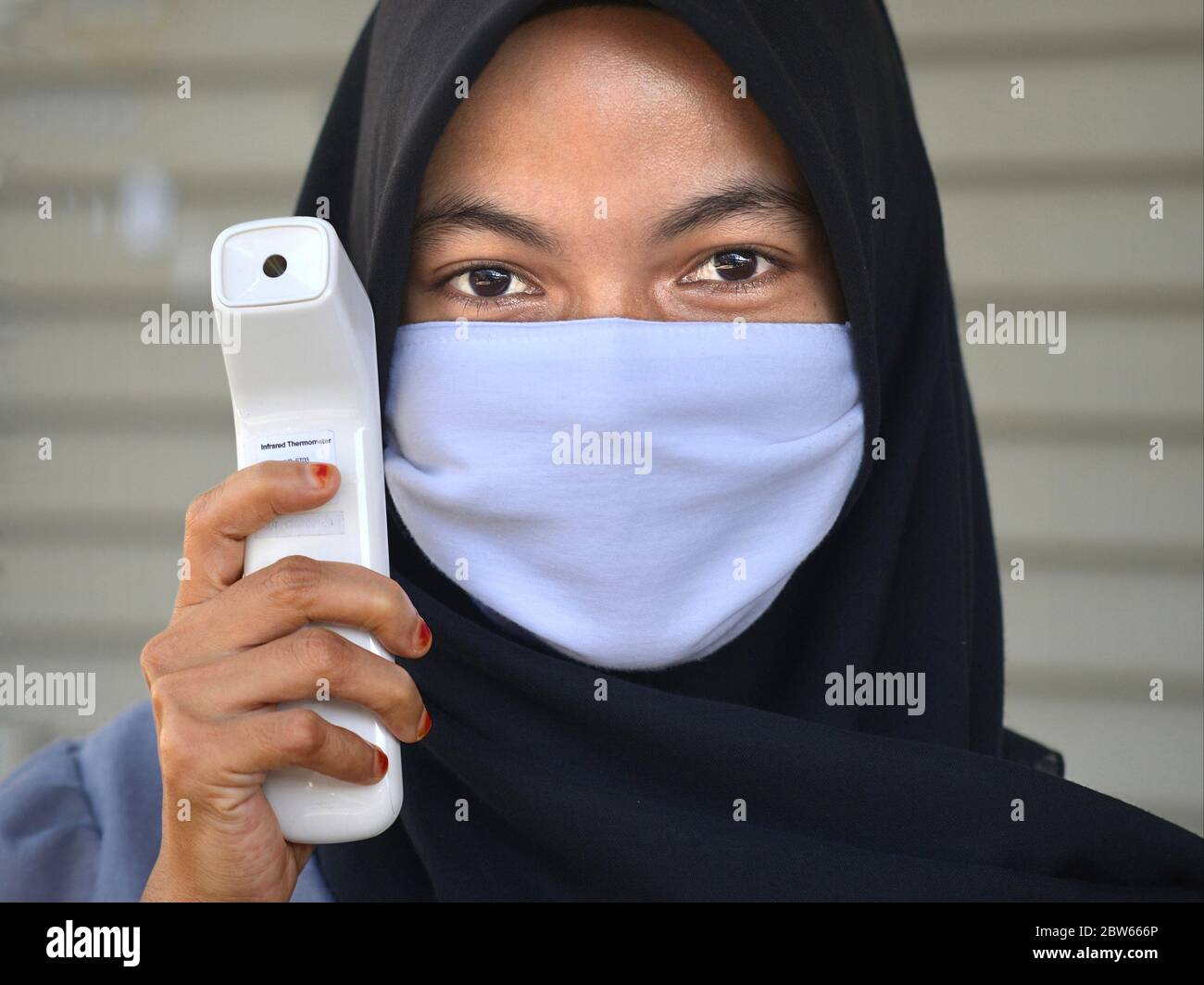 Malay girl wears a black traditional hijab with mouth-covering veil and poses for the camera with a modern infrared thermometer. Stock Photo