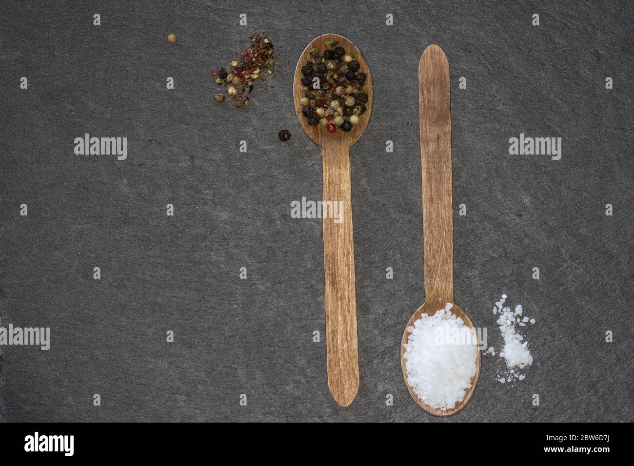 two wooden spoons with coarse salt and colored pepper on a dark slate plate Stock Photo