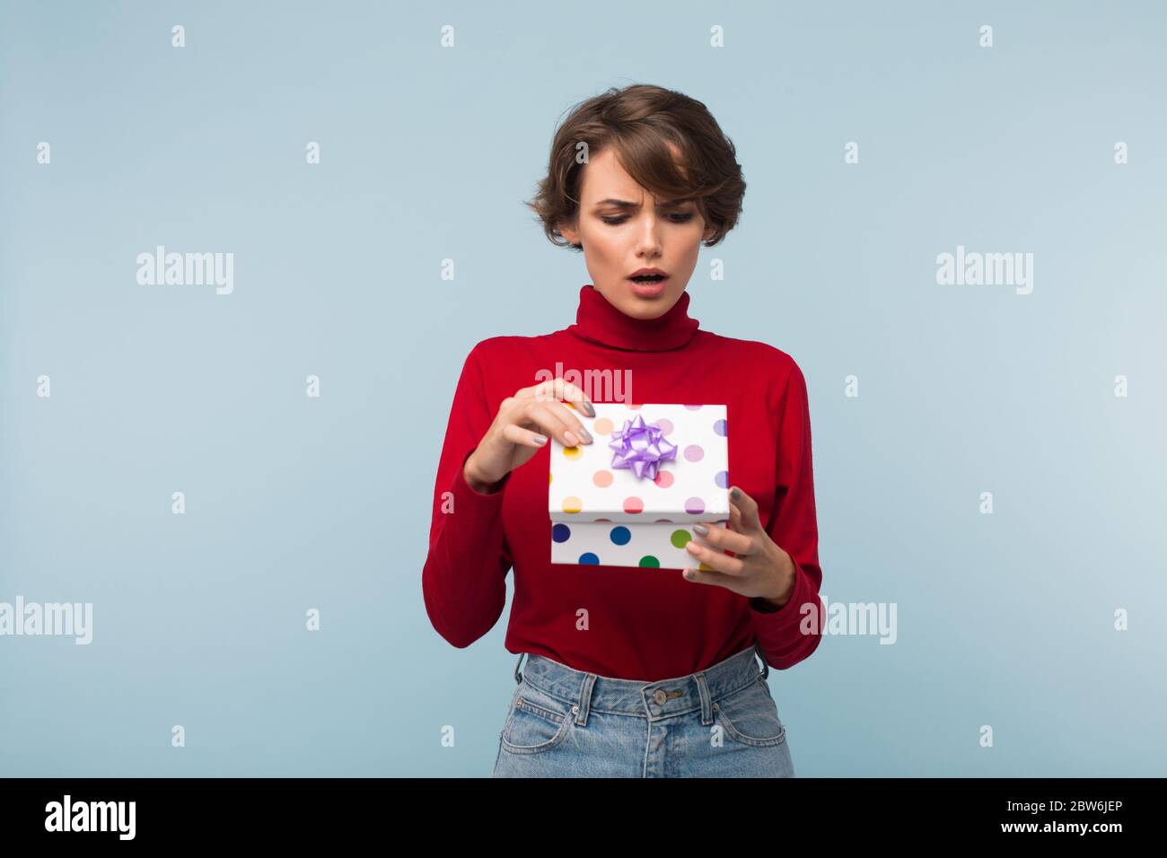 Young serious woman with dark short hair in red sweater sadly opening little present box over blue background isolated Stock Photo
