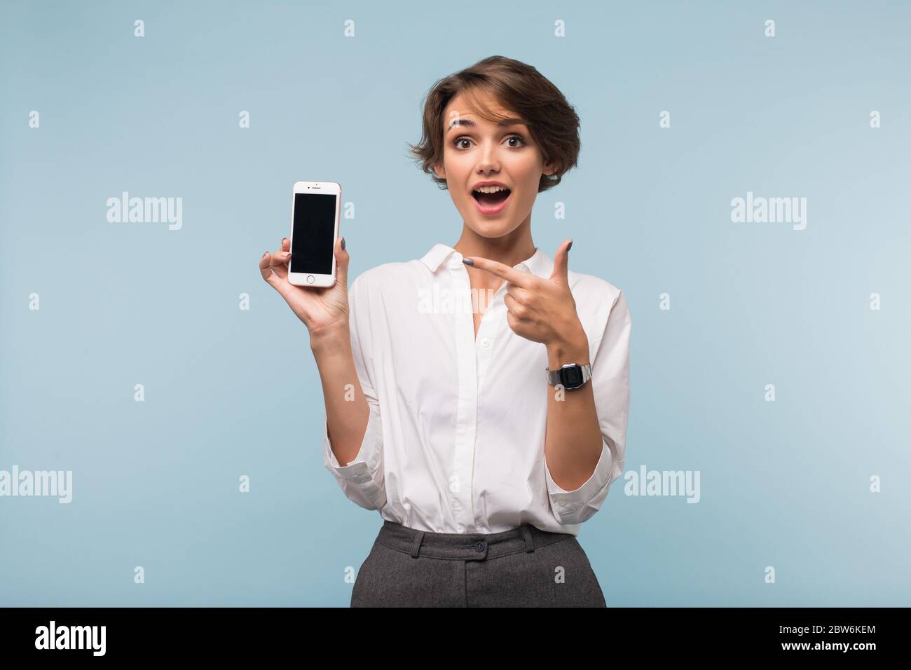 Young joyfull woman with dark short hair in white shirt amazedly showing cellphone on camera over blue background isolated Stock Photo