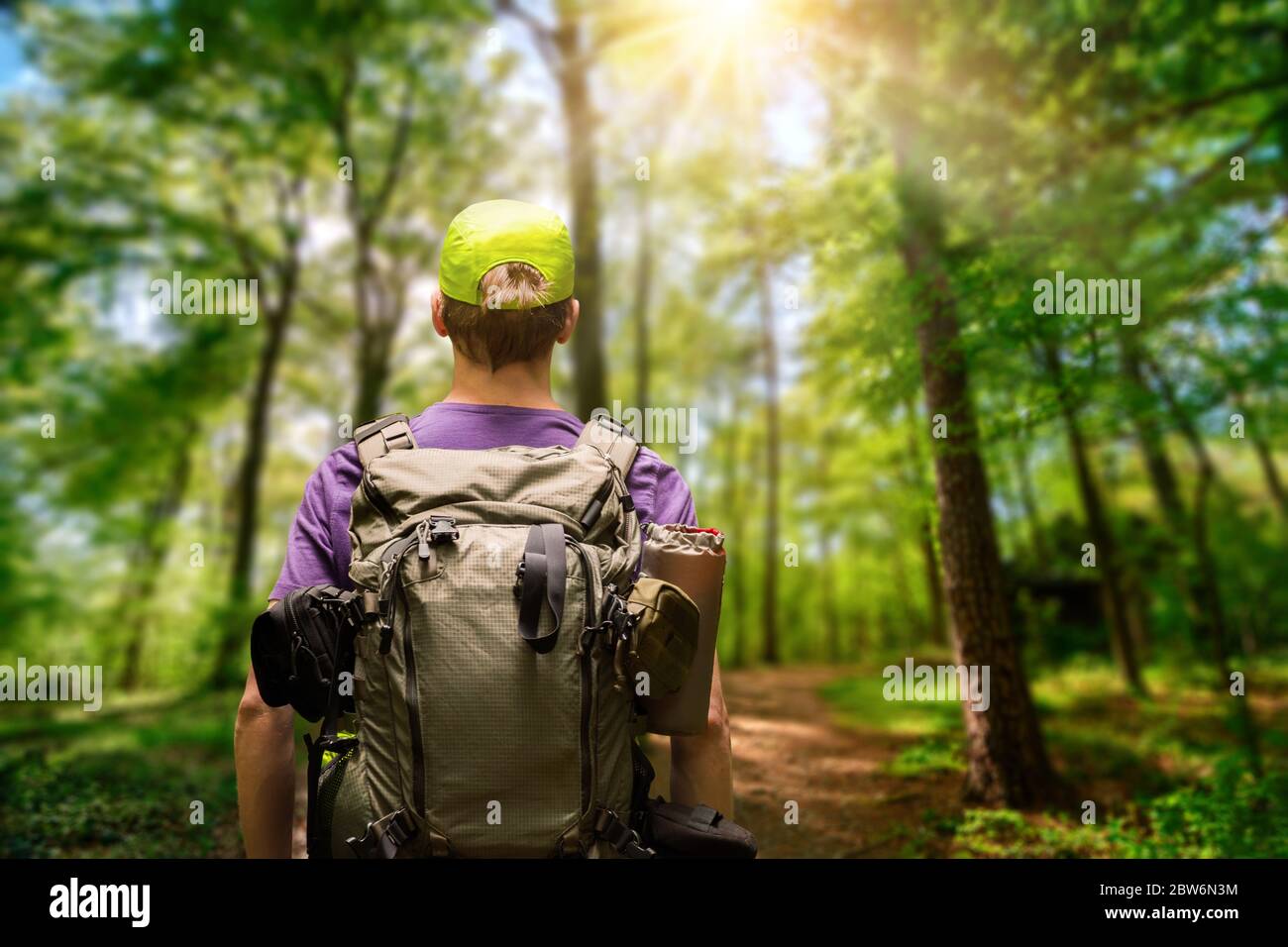 Male hiker with a backpack exploring a path in a green forest with the sun shining above, shallow focus Stock Photo