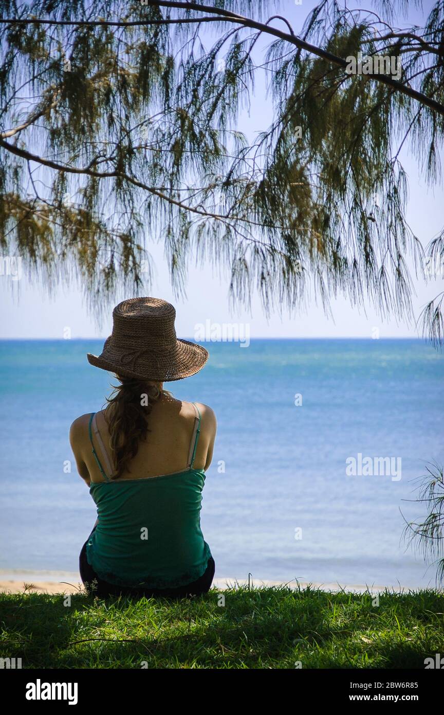 Female tourist sitting, framed under a Casuarina, on a grassed beachfront looking out on the Pacific Ocean at Palm Cove in Far North Queensland. Stock Photo