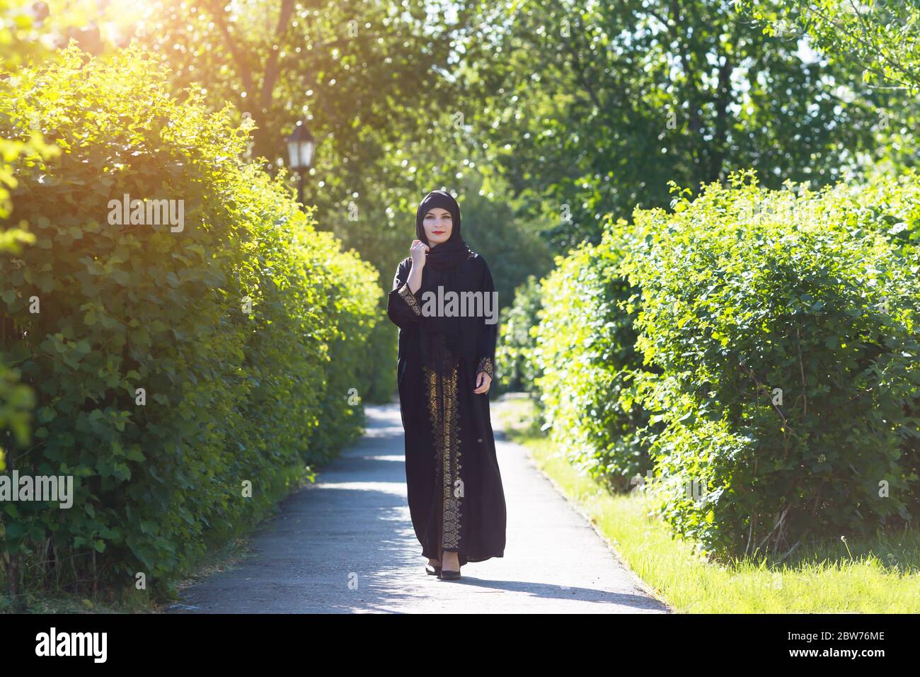 A girl in Islamic oriental clothes beautifully walks along the alley among the trees. Stock Photo