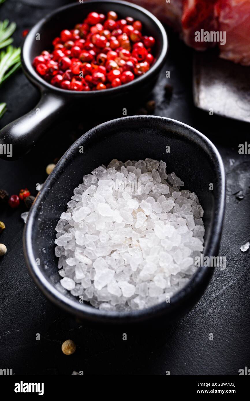 Sea Salt in black bowl and Rosemary herbs close up on black stone table with spices and raw meat near side view selective focus vertical. Stock Photo