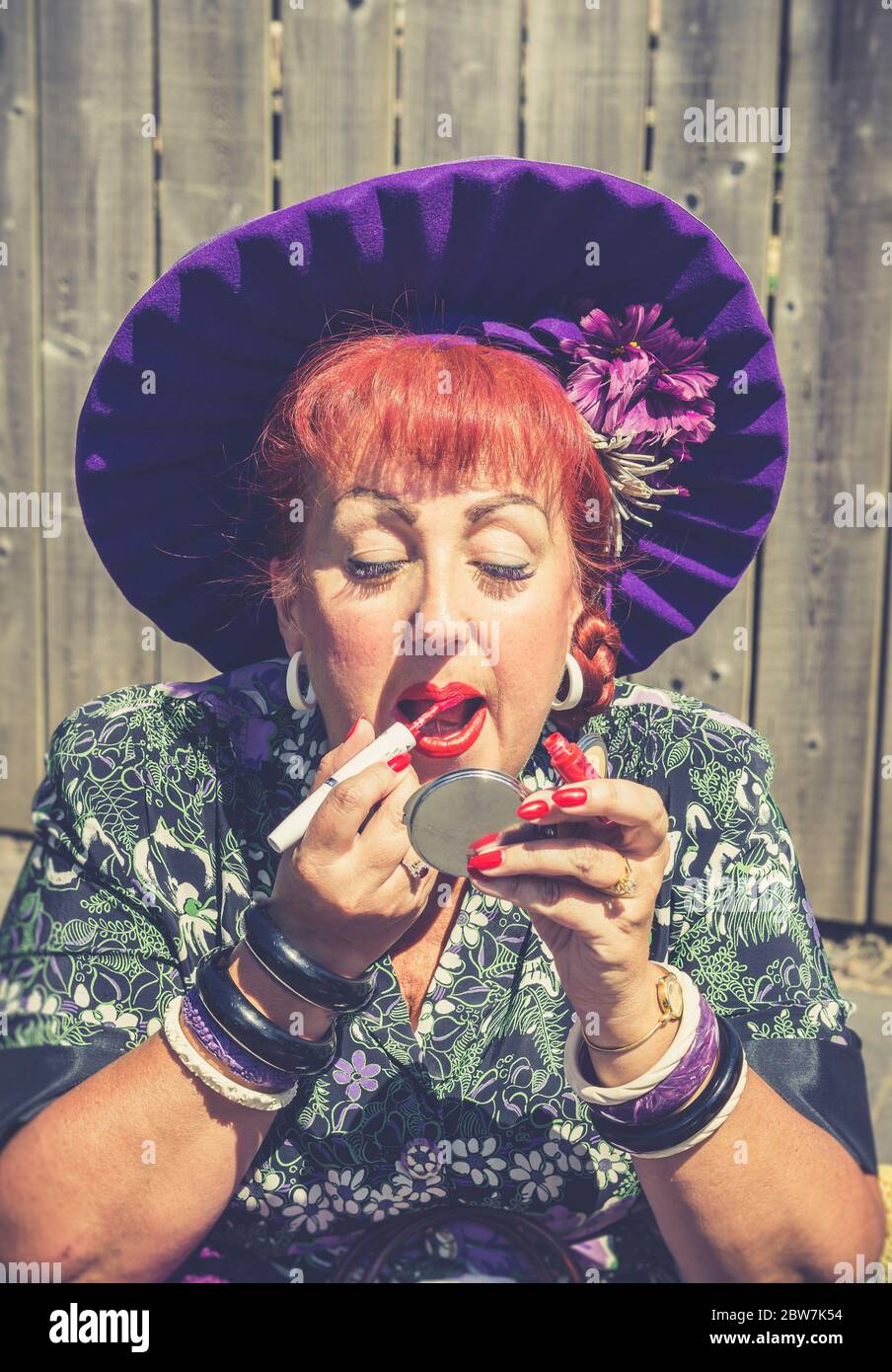 Close up of isolated woman in bid, purple hat putting on bright red lipstick, outdoors in sunshine at 1940s WW2 reenactment event, UK. Stock Photo