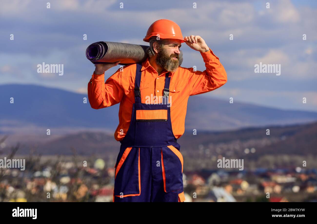 Roof installation. Man hard hat work outdoor landscape background. Building house. Apply plastic coatings membranes fiberglass or felt over sloped roofs before applying shingles. Roofer repair roof. Stock Photo