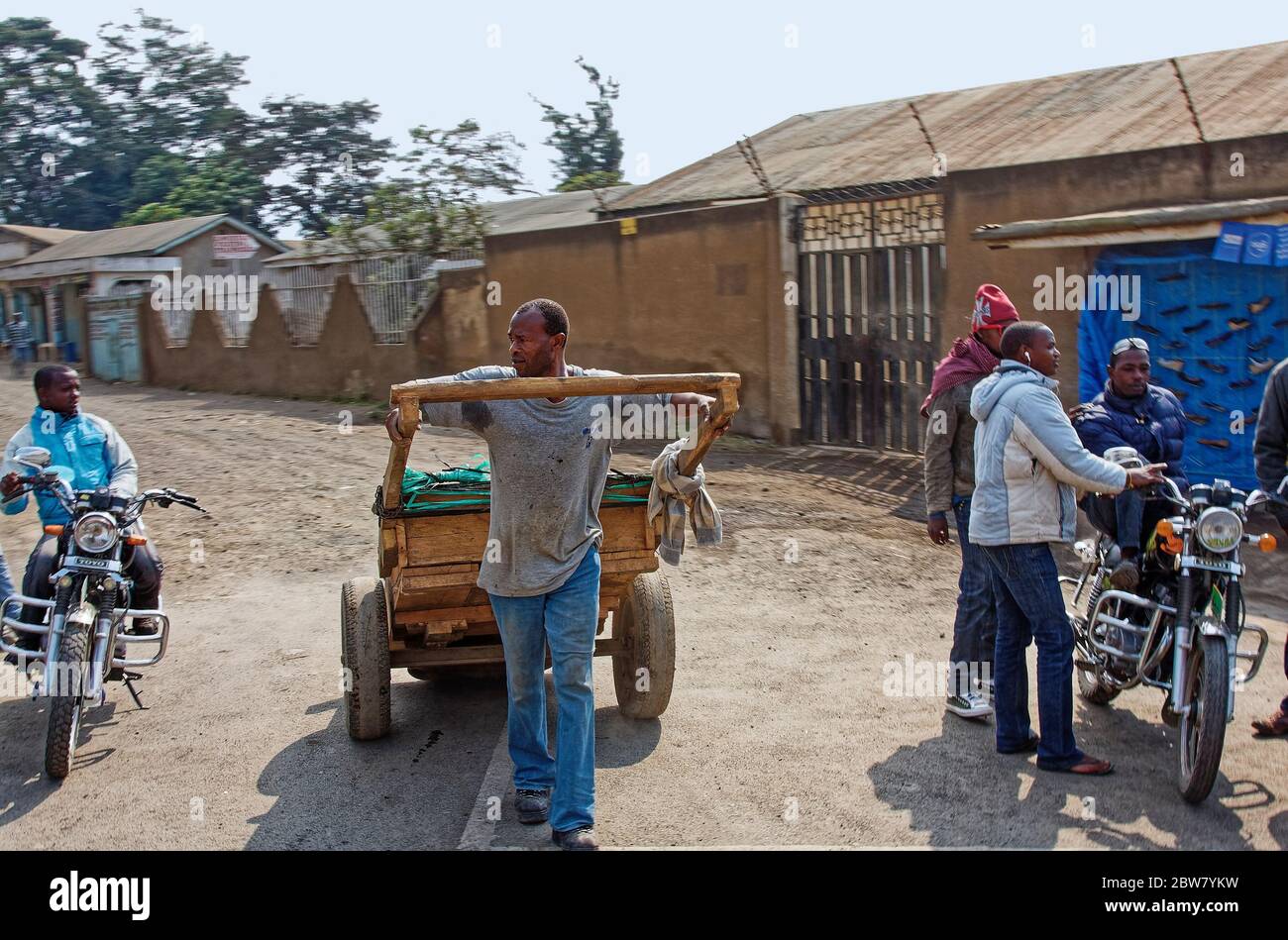 man pulling laarge cart, sweating, perspiration stains, working hard, job, people talking, motorcycles, shopping area, Tanzania; Africa Stock Photo