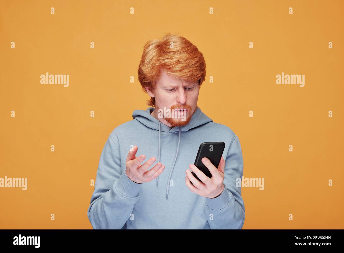 Confused or dissatisfied young man in hoodie talking to someone while looking at screen of smartphone over yellow background Stock Photo