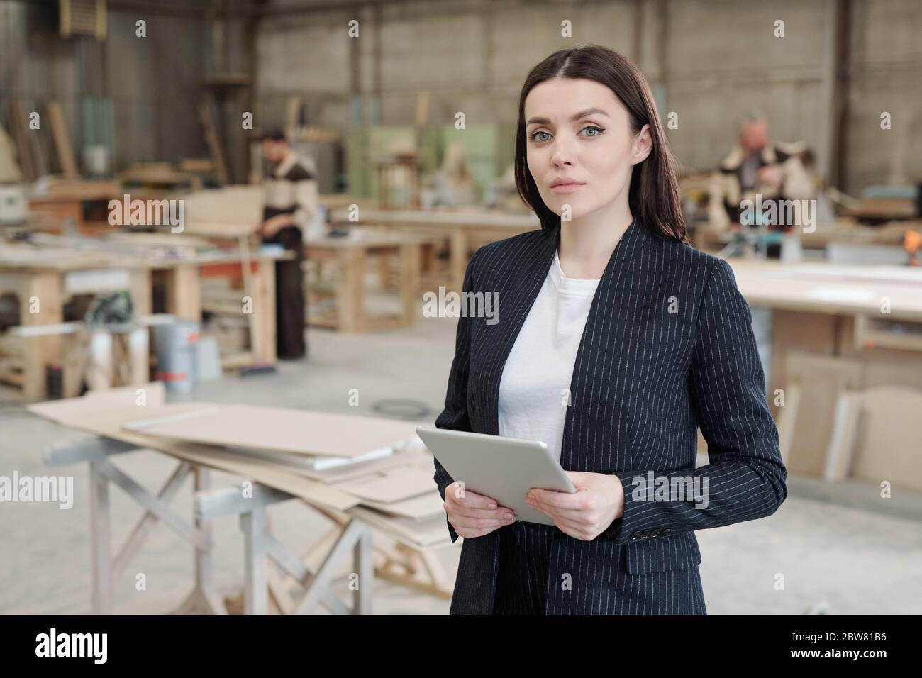 Young serious sales manager of furniture factory using touchpad in front of camera while standing against workshop interior and male workers Stock Photo