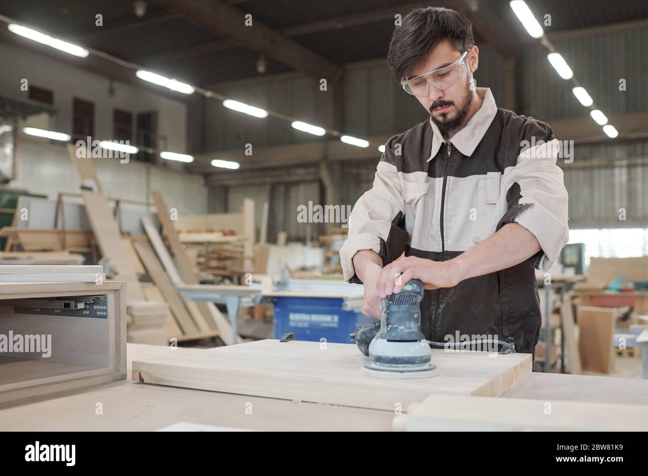Young man in workwear and protective eyeglasses bending over workbench
