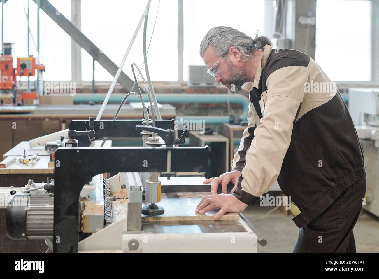 Mature engineer of furniture factory bending over industrial machine in