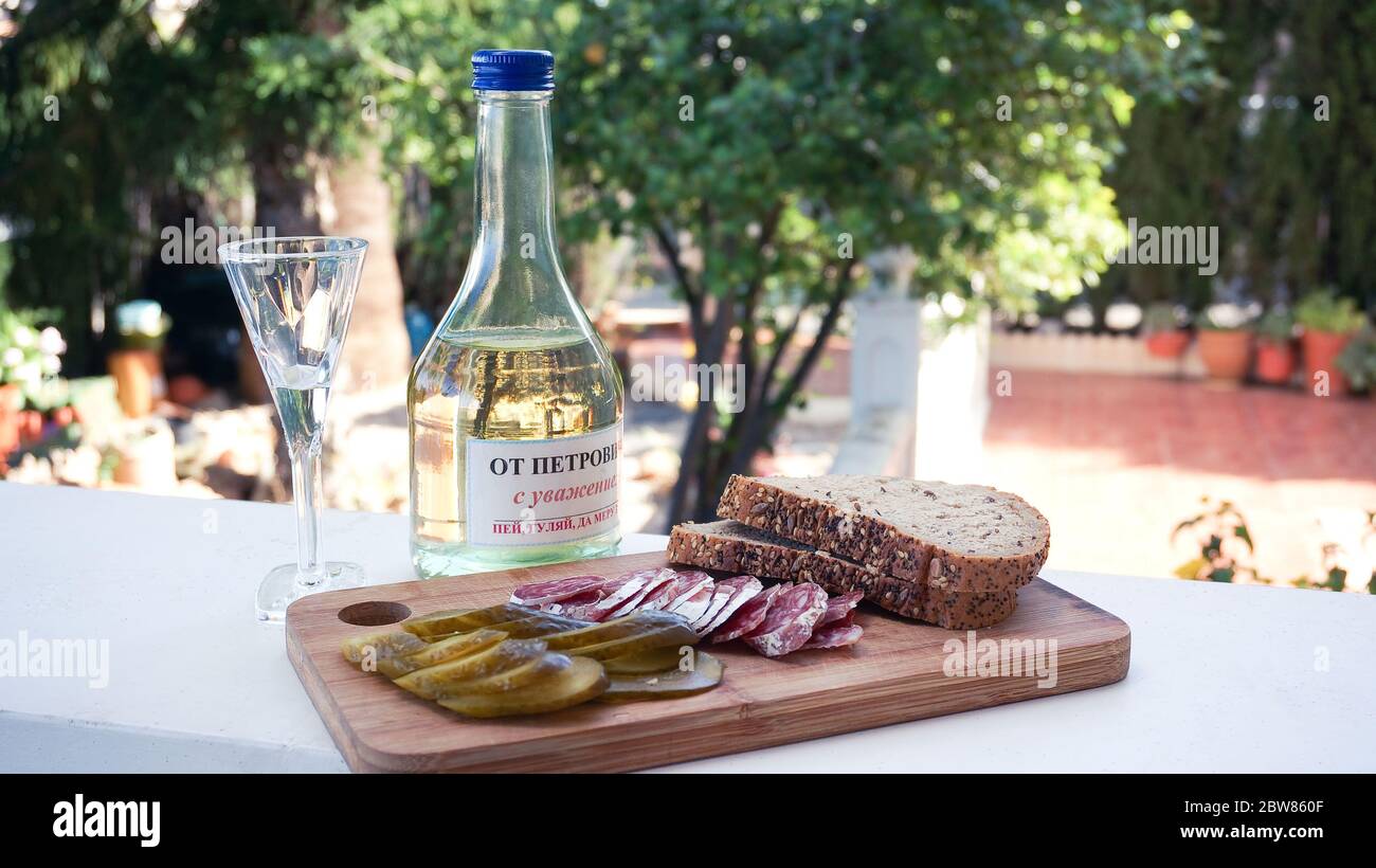 Homemade traditional russian vodka 'Samogon Regards From Petrovich' written on bottle label. Saussage, pickeles and bread on wooden board as a snack s Stock Photo
