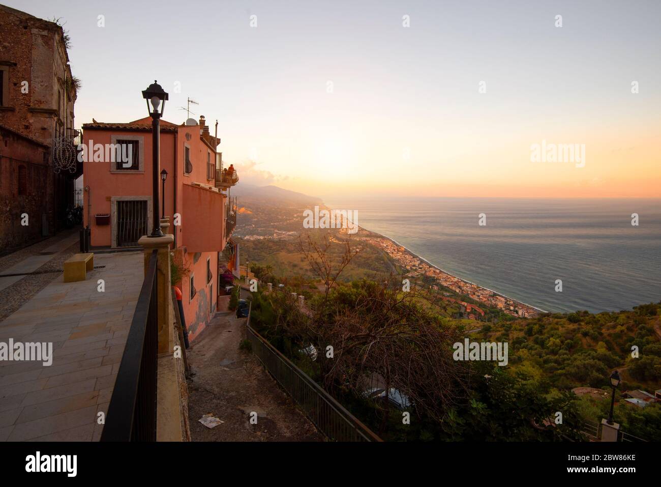 A beautiful glaring sunrise lighting up the red buildings in the foreground with a narrow street and lamp. To the right is a view of the coast Stock Photo