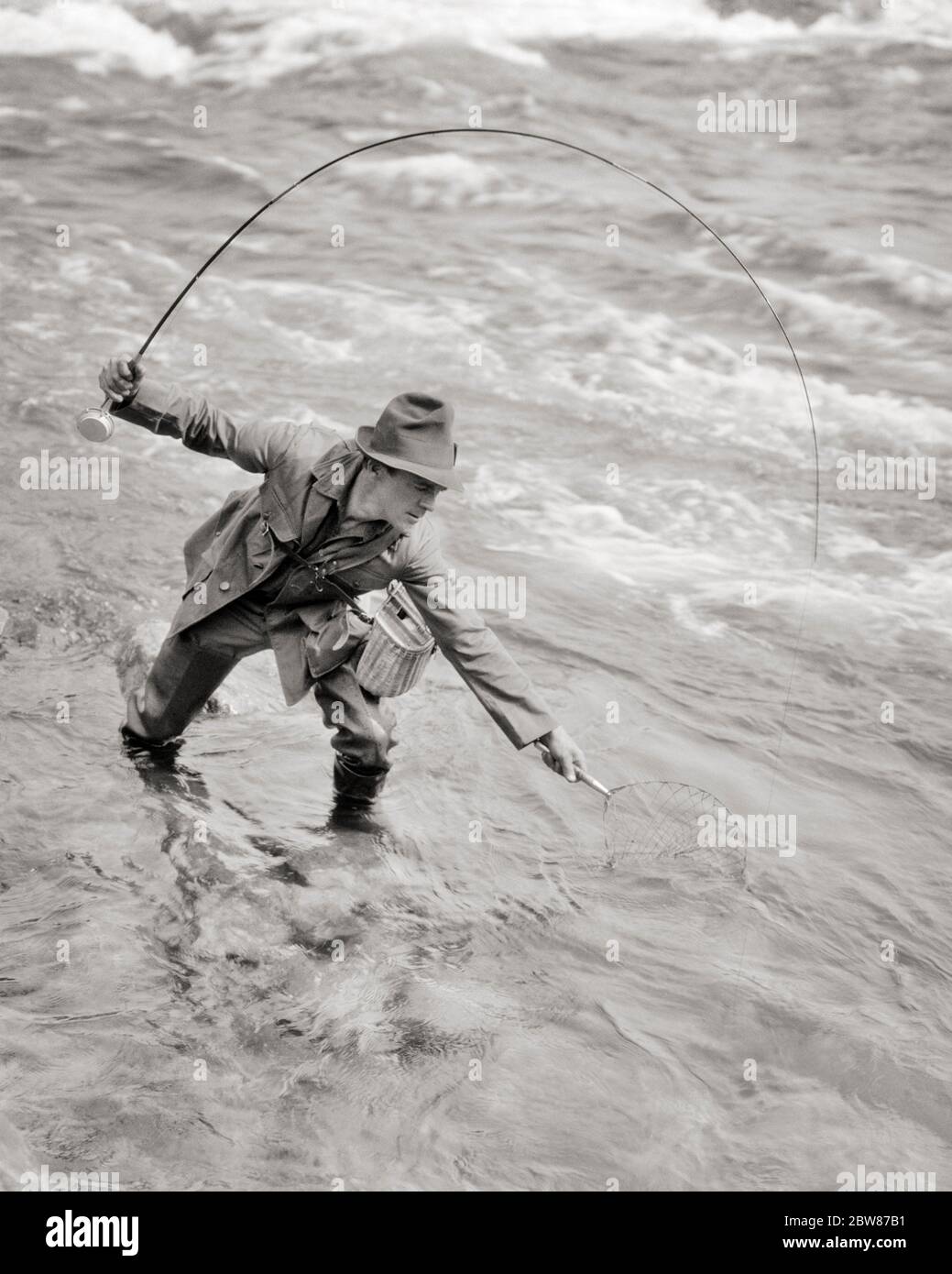 1920s MAN FISHERMAN STANDING IN STREAM BENDING OVER WITH NET TO CATCH FISH  HOOKED ON HIS LINE - a863 HAR001 HARS RURAL HEALTHINESS NATURE COPY SPACE  CATCH FULL-LENGTH PERSONS STREAM MALES ATHLETIC
