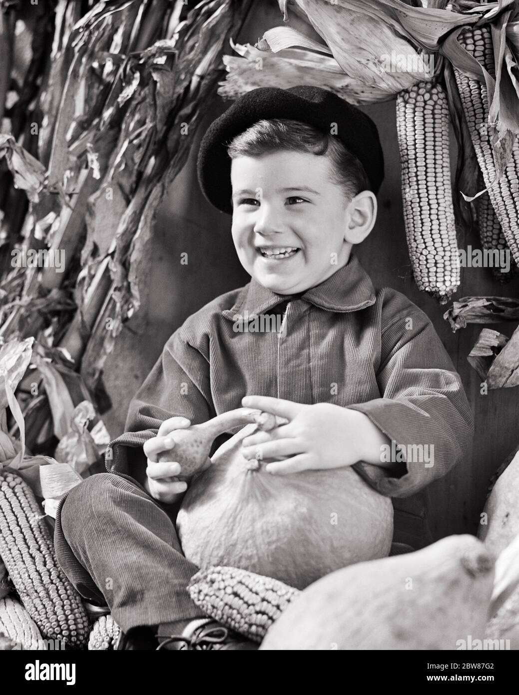 1940s 1950s SMALL BOY SMILING SITTING WITH AUTUMN PUMPKIN GOURDS EARS OF DRIED CORN WEARING CORDUROY JACKET AND TROUSERS - c1743 HAR001 HARS CONFIDENCE AGRICULTURE B&W BRUNETTE HAPPINESS CHEERFUL TROUSERS DRIED AND CORDUROY FALL SEASON PRIDE SMILES JOYFUL GOURDS JUVENILES AUTUMNAL BLACK AND WHITE CAUCASIAN ETHNICITY FALL FOLIAGE HAR001 OLD FASHIONED Stock Photo