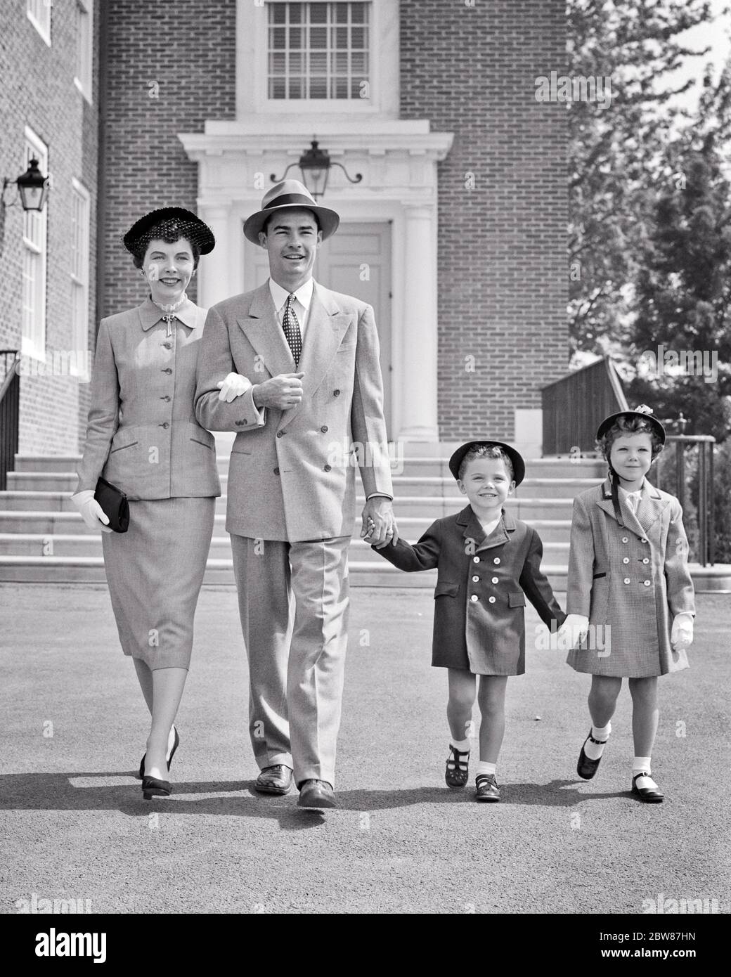 1950s FAMILY OF FOUR DRESSED IN THEIR SUNDAY BEST CLOTHES WALKING AWAY FROM CHURCH BUILDING SMILING MOTHER FATHER BOY GIRL - c4683 HAR001 HARS SUBURBAN SPRING MOTHERS OLD TIME NOSTALGIA BROTHER OLD FASHION SISTER JUVENILE STYLE COMMUNICATION TEAMWORK BEST PLEASED FAMILIES JOY LIFESTYLE SATISFACTION RELIGION FEMALES EASTER MARRIED SUNDAY BROTHERS SPOUSE HUSBANDS COATS HEALTHINESS COPY SPACE FRIENDSHIP FULL-LENGTH LADIES PERSONS INSPIRATION AWAY FESTIVAL MALES CHRISTIAN SIBLINGS SPIRITUALITY CONFIDENCE SISTERS FATHERS B&W PARTNER SUIT AND TIE HAPPINESS CHEERFUL LEISURE RELIGIOUS CHRISTIANITY Stock Photo