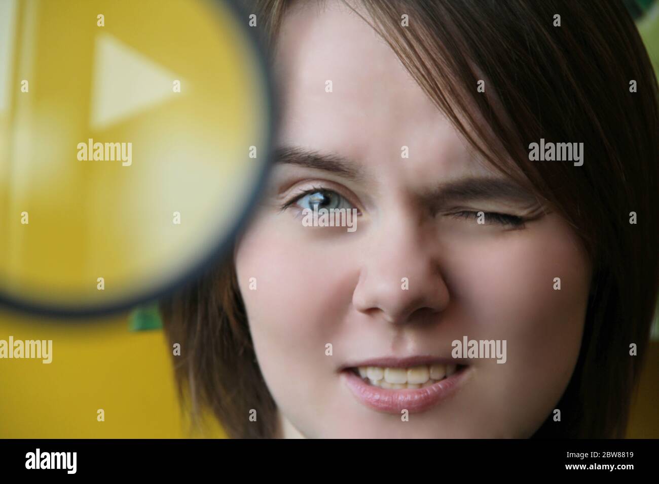 A beautiful young girl looks through a magnifying glass squinting one eye. Face close-up.  Stock Photo