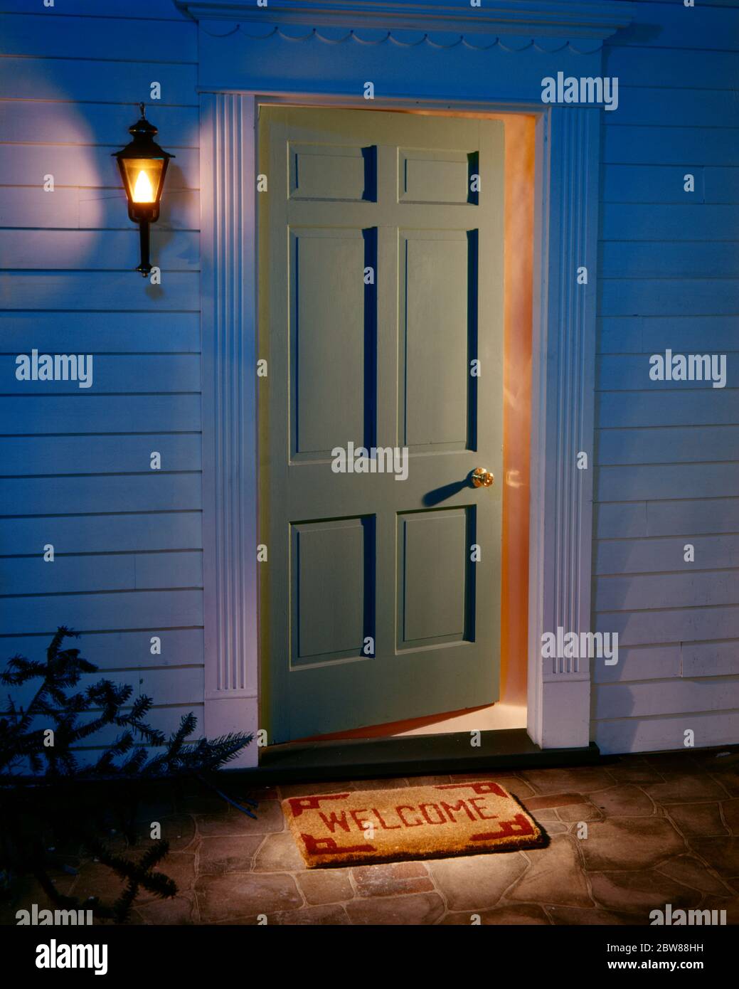 1970s OPEN FRONT HOME DOORWAY AT NIGHT WITH WELCOME MAT LIGHTS ON INSIDE AND LANTERN ON SIDE OF HOUSE  - kd2844 PHT001 HARS AT OF ON HOMES CONCEPT CONCEPTUAL RESIDENCE SYMBOLIC CONCEPTS OLD FASHIONED REPRESENTATION Stock Photo