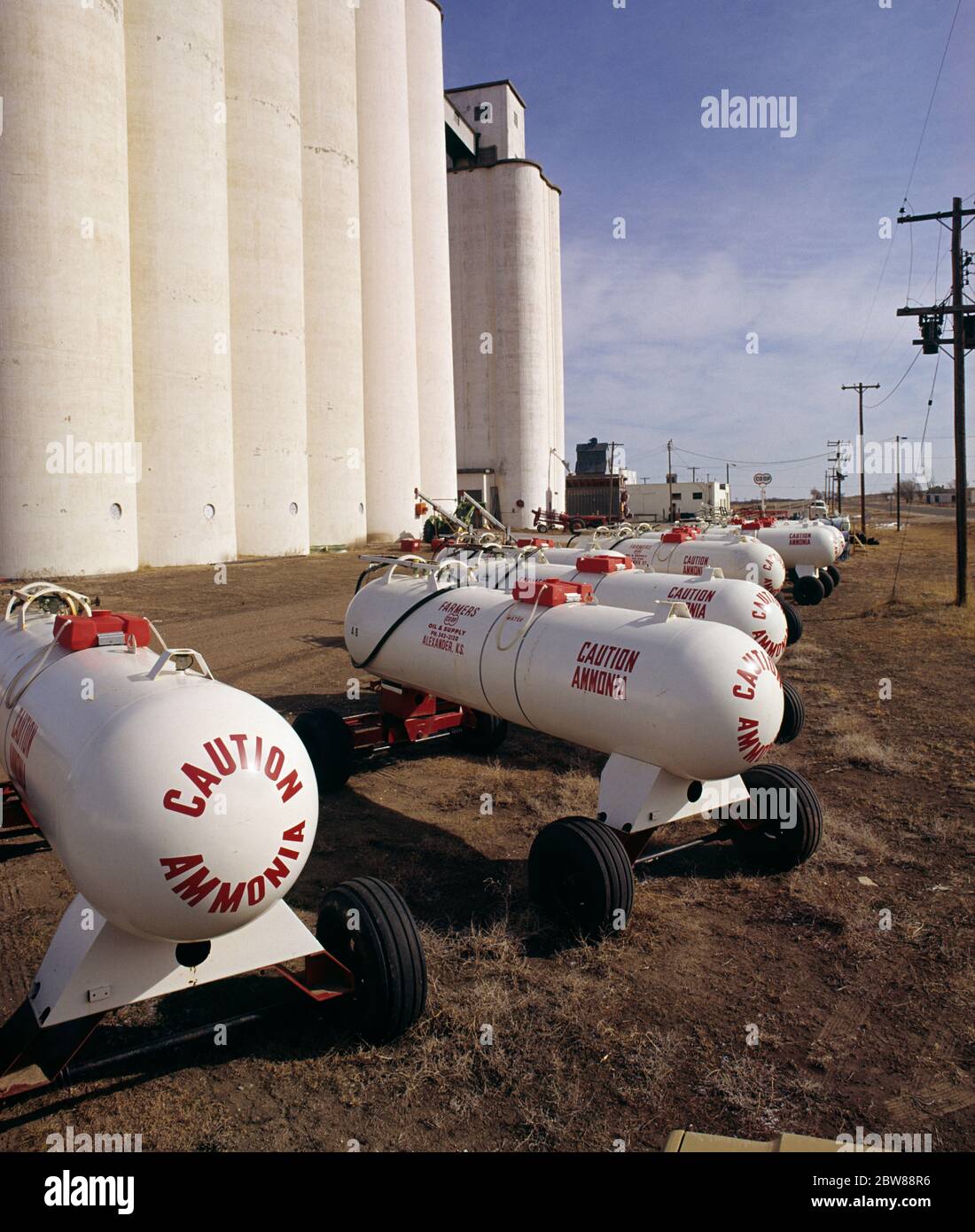 1980s FARMERS COOP WHITE GRAIN SILOS RED AND WHITE TANKS OF ANHYDROUS AMMONIA FOREGROUND CAUTION