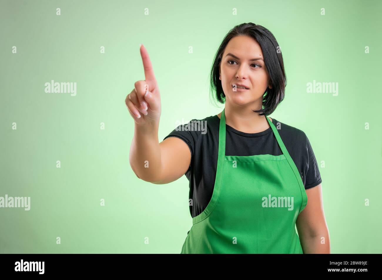 Female cook with green apron and black t-shirt, presses a virtual button isolated on green background Stock Photo