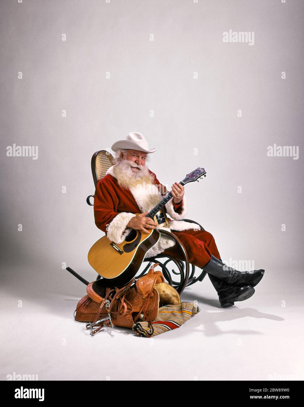 1980s COWBOY SANTA CLAUS LOOKING AT CAMERA SITTING IN BENTWOOD ROCKER BESIDE WESTERN SADDLE PLAYING GUITAR WEARING WHITE HAT - kx11156 PHT001 HARS NOSTALGIC SYMBOL SUBURBAN URBAN XMAS COLOR OLD TIME BUSY NOSTALGIA OLD FASHION BEARD 1 FACIAL STYLE COMMUNICATION EVE PEACE ROCKING JOY LIFESTYLE MUSICIAN JOBS STUDIO SHOT ROCKER RURAL GROWNUP CLAUS HOME LIFE COPY SPACE FULL-LENGTH ICON PERSONS SADDLE WISE GROWN-UP CARING CHARACTER MALES WESTERN ENTERTAINMENT SPIRITUALITY SENIOR MAN SAINT SENIOR ADULT WINTERTIME EYE CONTACT WINTER SEASON MUSTACHE PERFORMING ARTS ICONS HAIRS OCCUPATION HAPPINESS Stock Photo