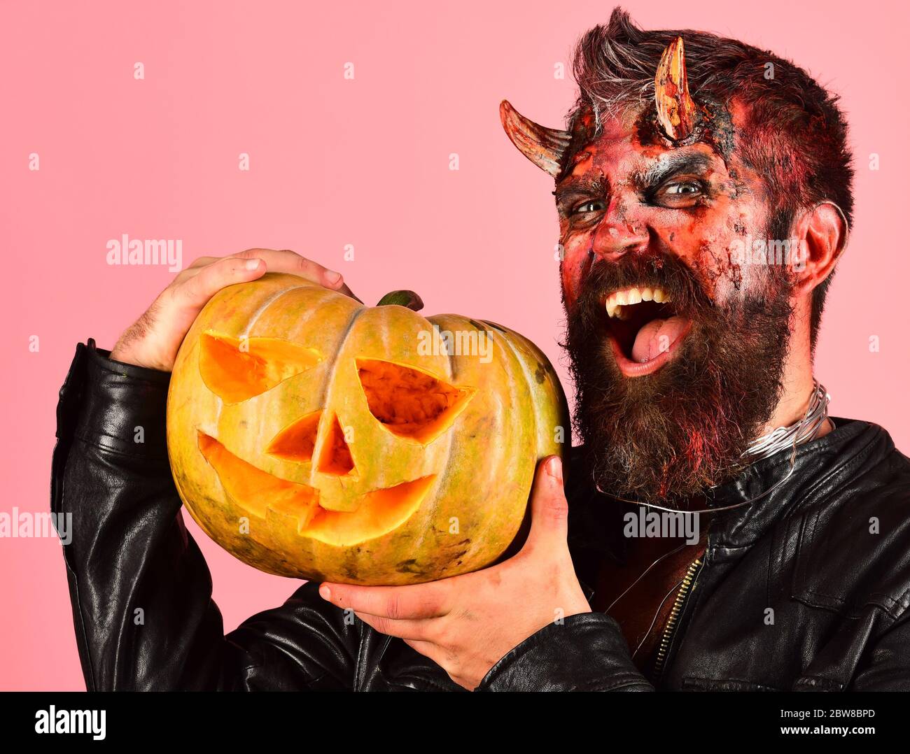 Man wearing scary makeup holds pumpkin on pink background. Halloween party  concept. Devil or monster with October decorations. Demon with horns and  evil smile holds carved jack o lantern Stock Photo -