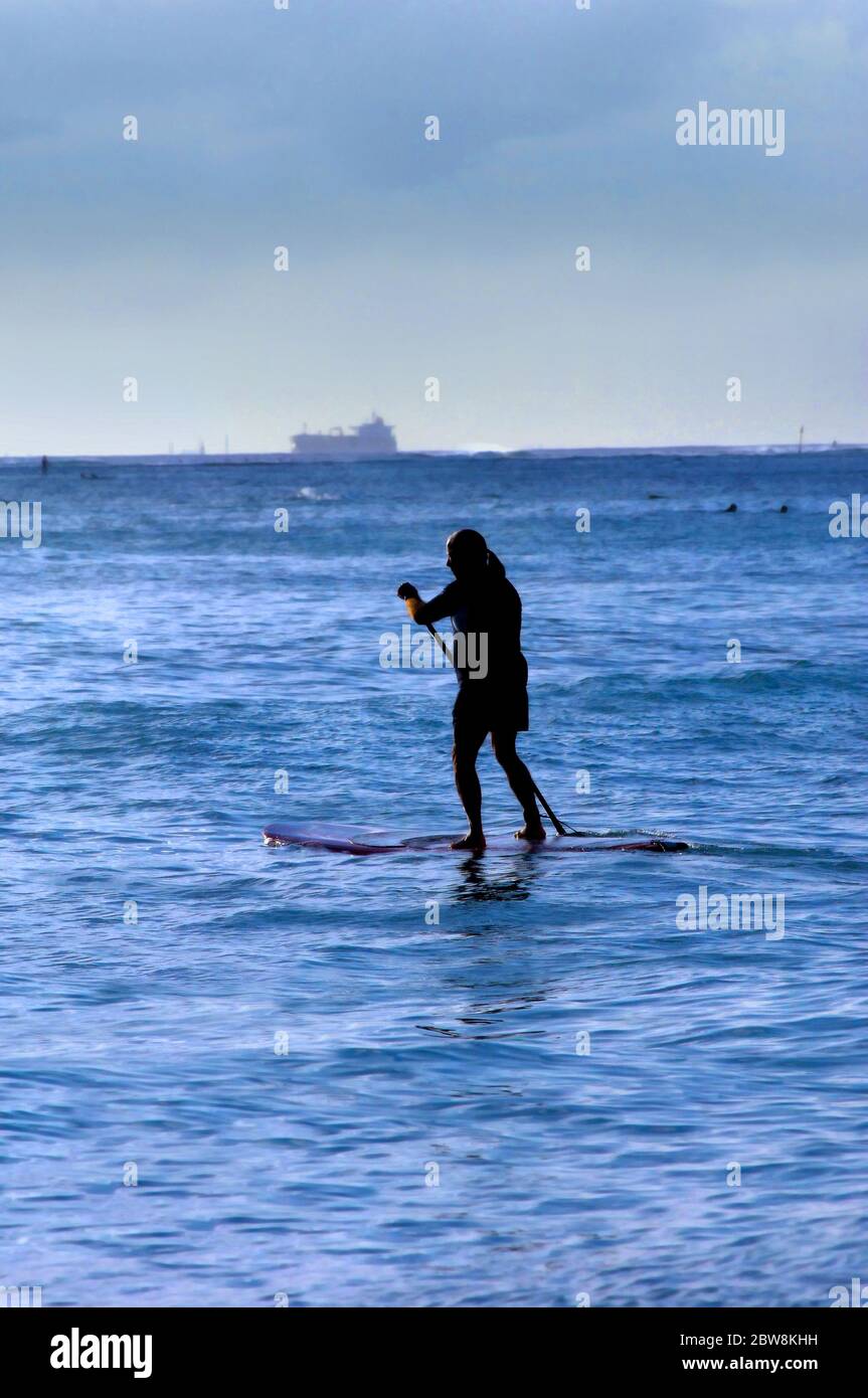 Older man is silhouetted as he paddle his boards across Waikiki Bay.  Ship is leaving port in background.  Evening light silhouettes paddler. Stock Photo