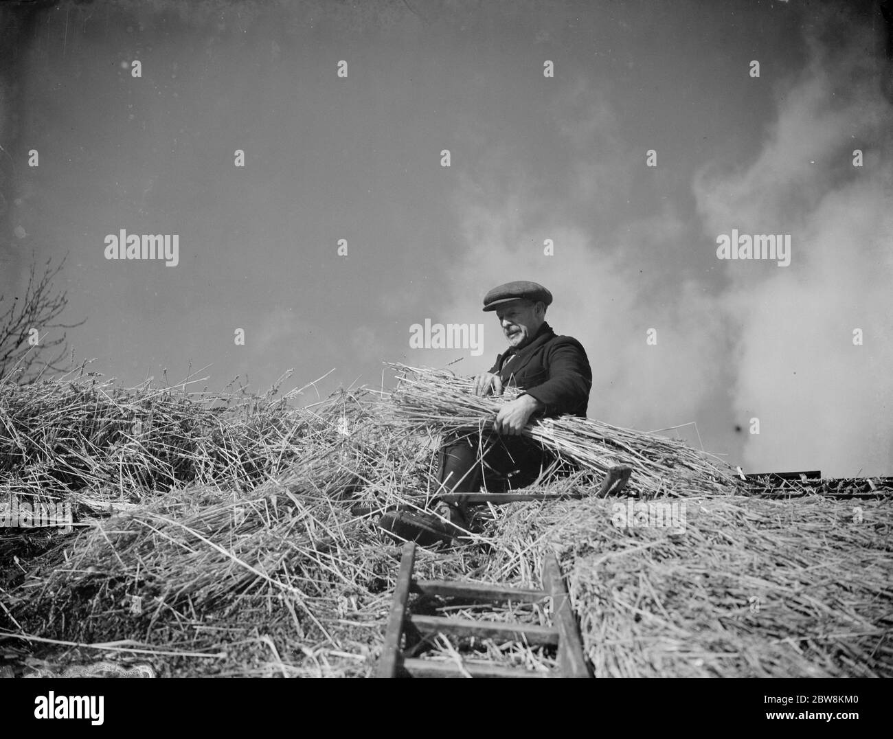 A man with a bundle of thatch in his arms works on the thatching on the roof of a cottage in Orpington . 1935 . Stock Photo