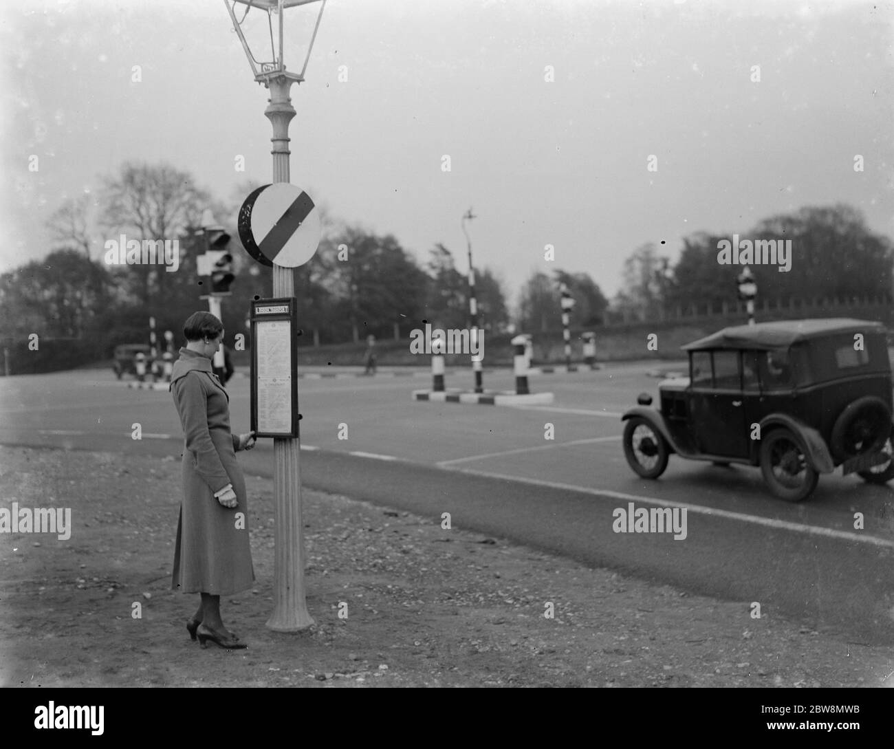A young woman studies the bus timetable at a traffic cross roads with traffic signs . 1935 Stock Photo