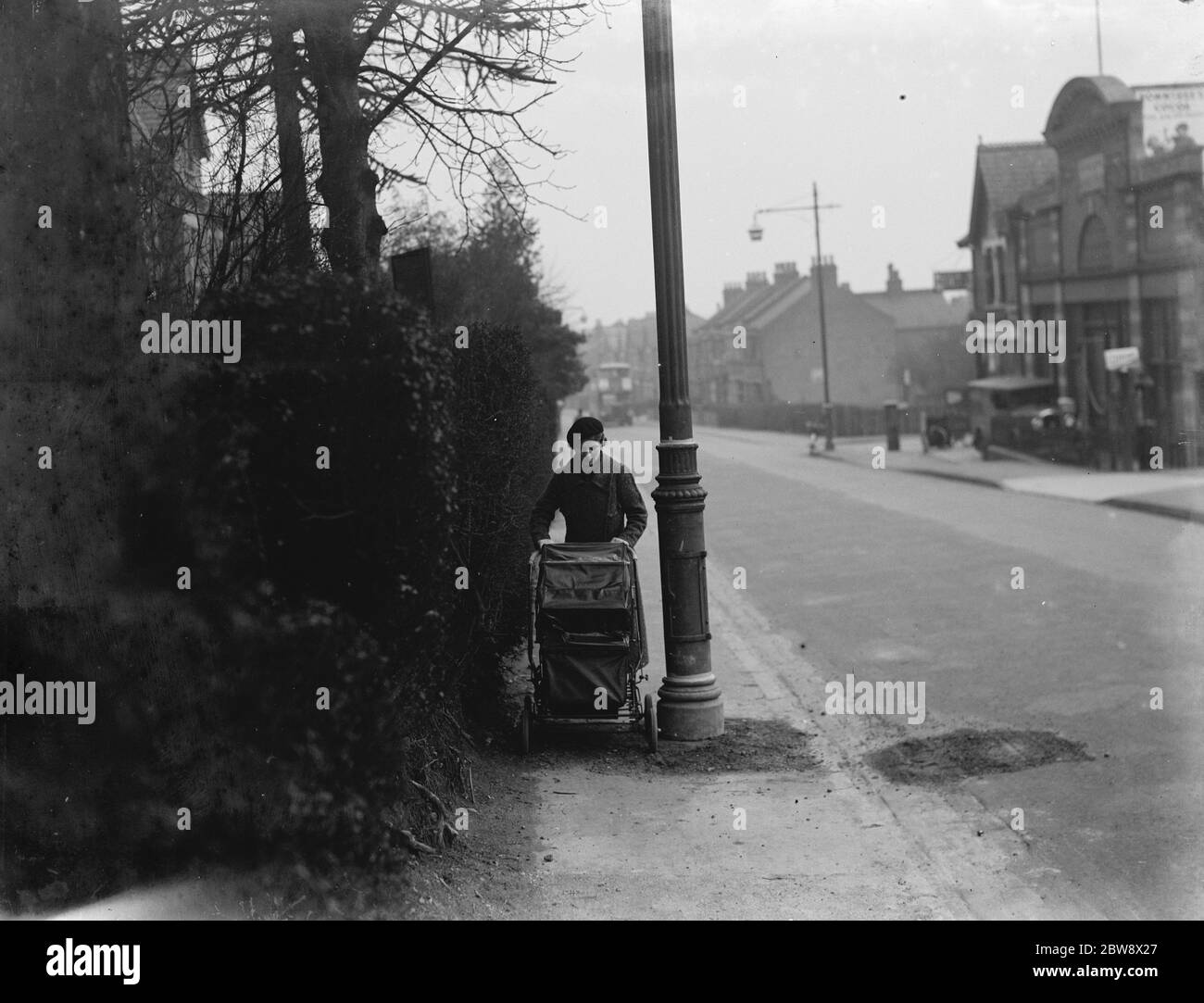 A woman pushes a pram past a street light which is shrouded by branches in Sidcup , Kent . 1937 Stock Photo