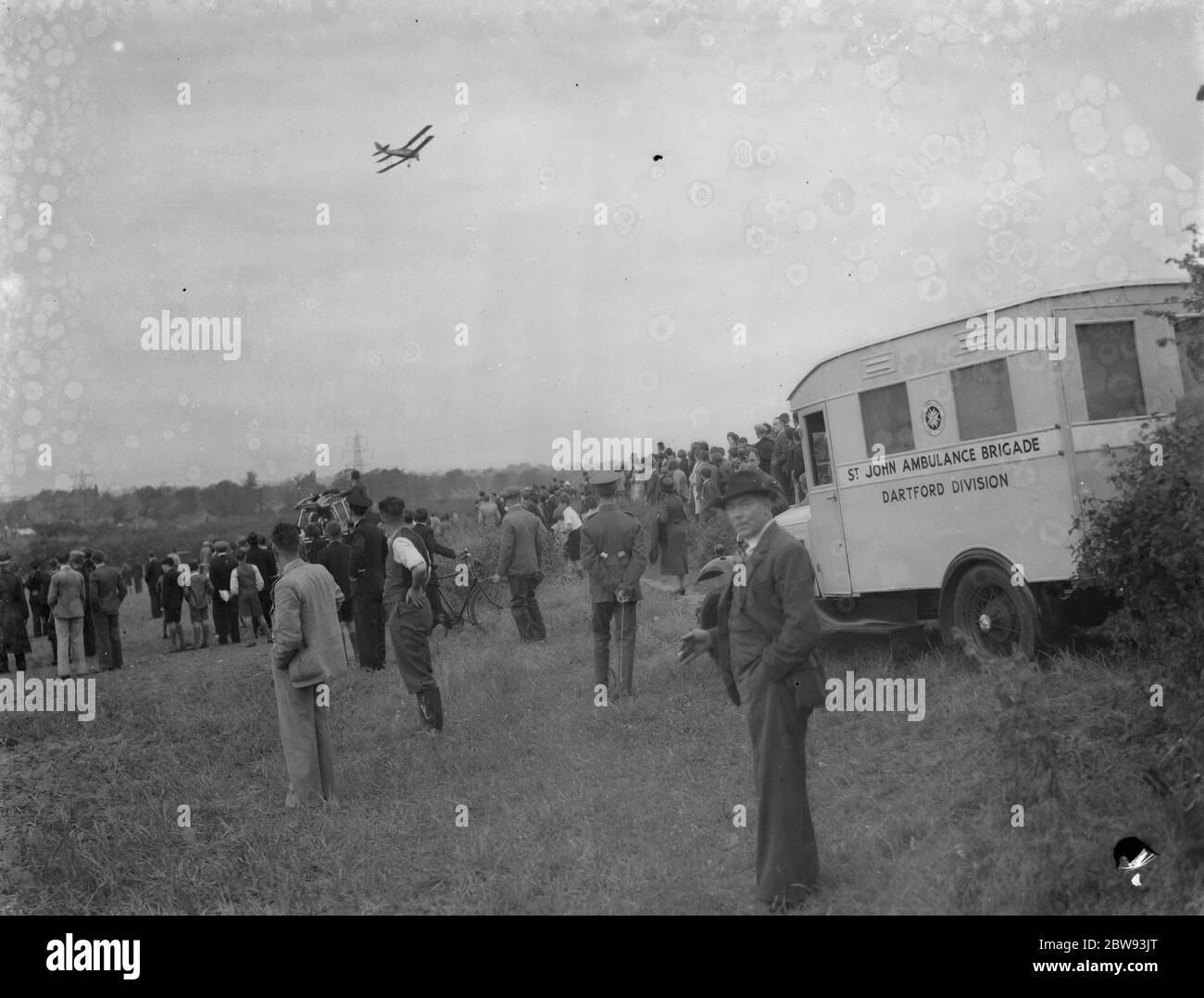 An ambulance at the scene where a de Havilland Tiger Moth aircraft crashed near Wilmington , Kent . Another de Havilland Tiger Moth can be seen circling overhead . 1939 Stock Photo