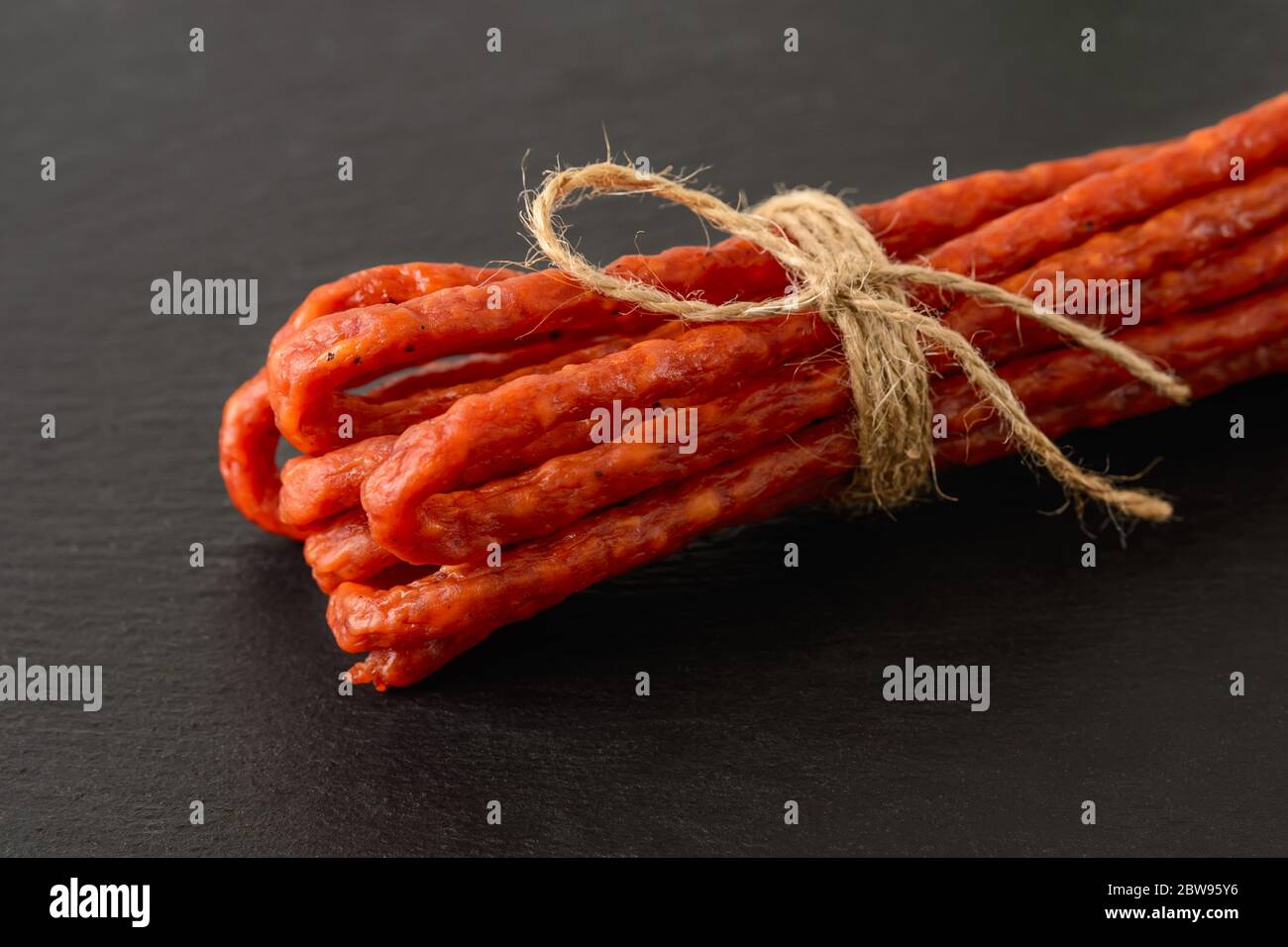 Close-up of thin dry smoked sausage kabanos or cabanossi on a black stone serving board. Traditional polish meat delicacy. Front view. Stock Photo