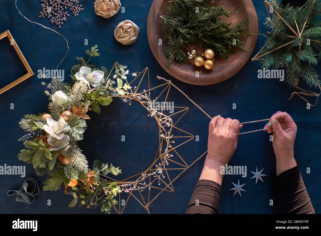 Making of decorative Christmas wreath on classic blue linen. Woman hands with handmade wreath. Xmas decorations, golden trinkets, decorative anemone f Stock Photo