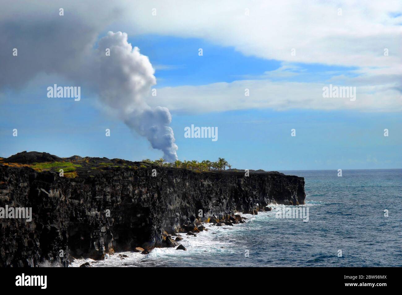 Vog billows from the active volcano on the Big Island of Hawaii.  Palm trees cling to the remaining area not yet covered by the flowing lava. Stock Photo