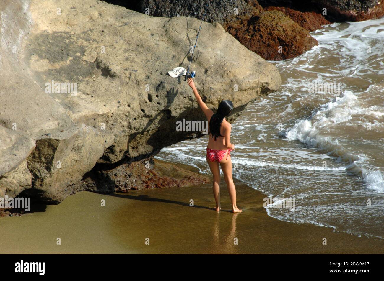 Female tourist reaches for her rod and reel on Green Sand Beach on the Big Island of Hawaii.  She has on a red print bikini. Stock Photo