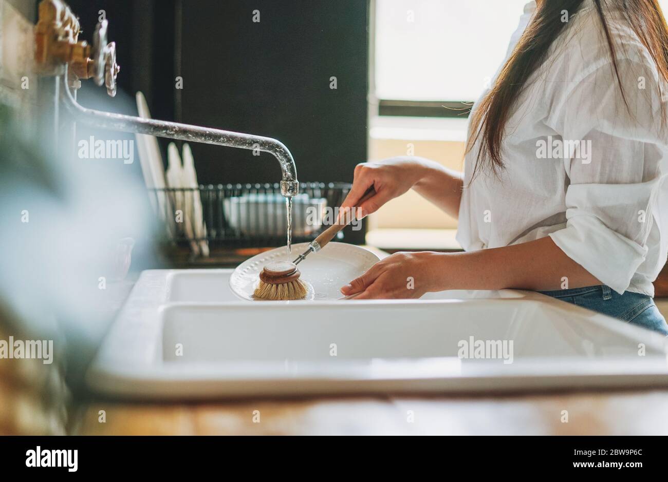 Young woman washes dishes with wooden brush with natural bristles at window in kitchen. Zero waste concept Stock Photo