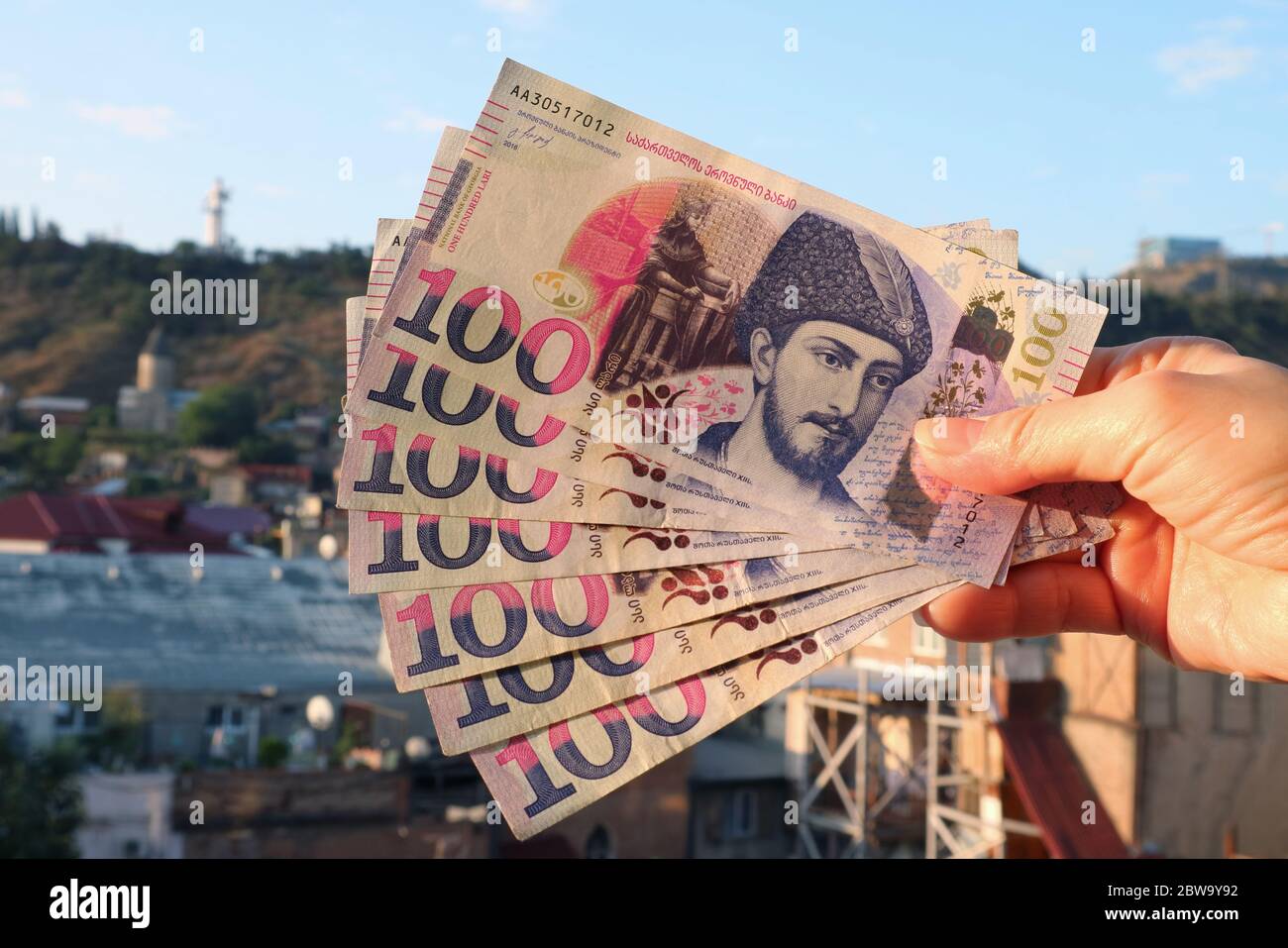 Hand Holding Bunch of 100 Georgian Lari banknotes in Obverse Side with Blurry Tbilisi Aerial View in the Backdrop Stock Photo