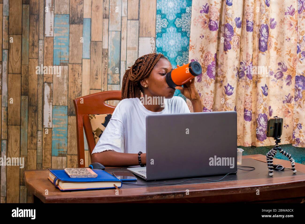 young black woman sitting at home, vlogging with her smartphone while working on her laptop Stock Photo