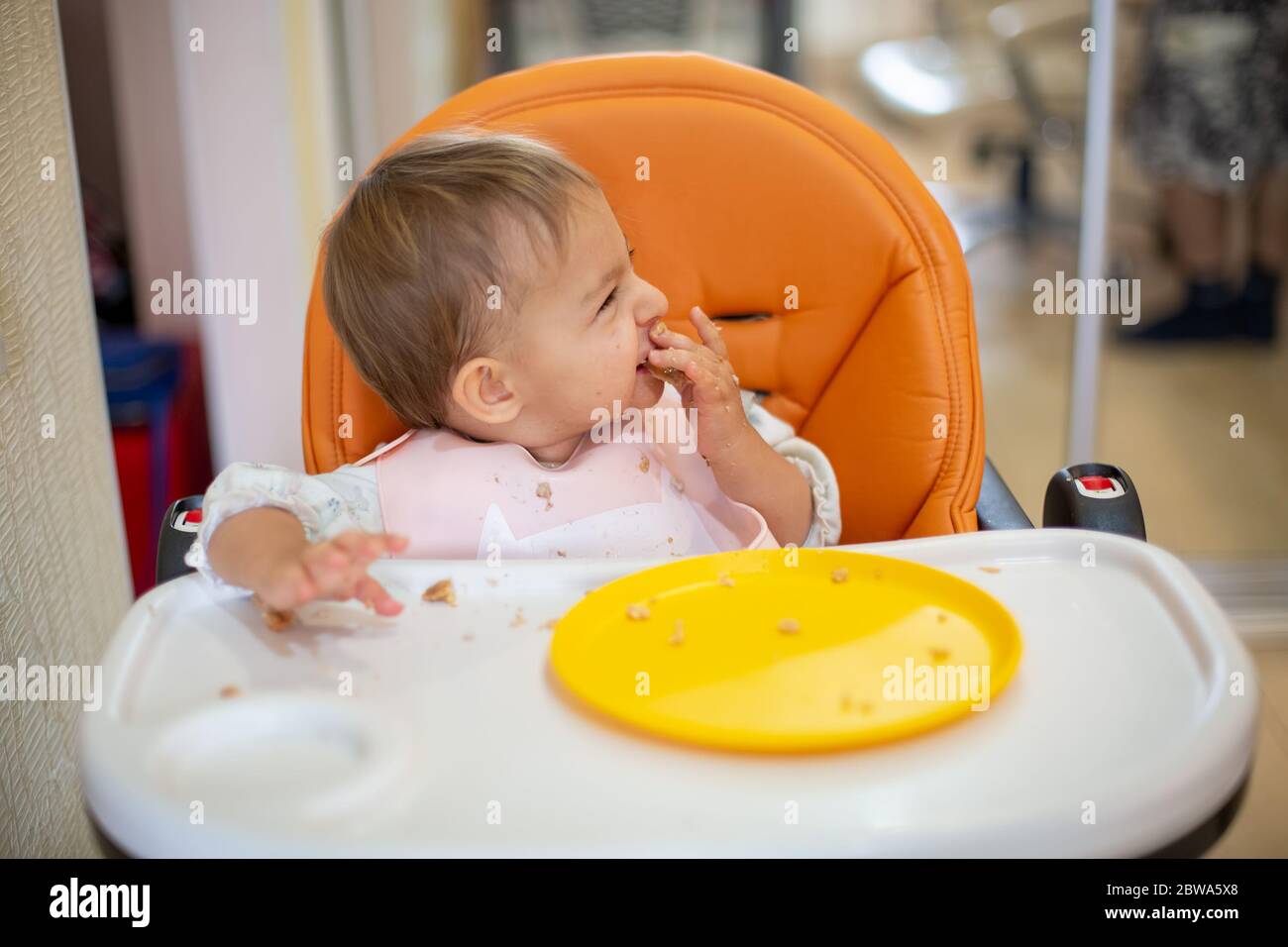 cute baby sits in an orange baby chair with a table with dirty hands and face, looks away and laughs. on table are crumbs and an orange plate. close Stock Photo