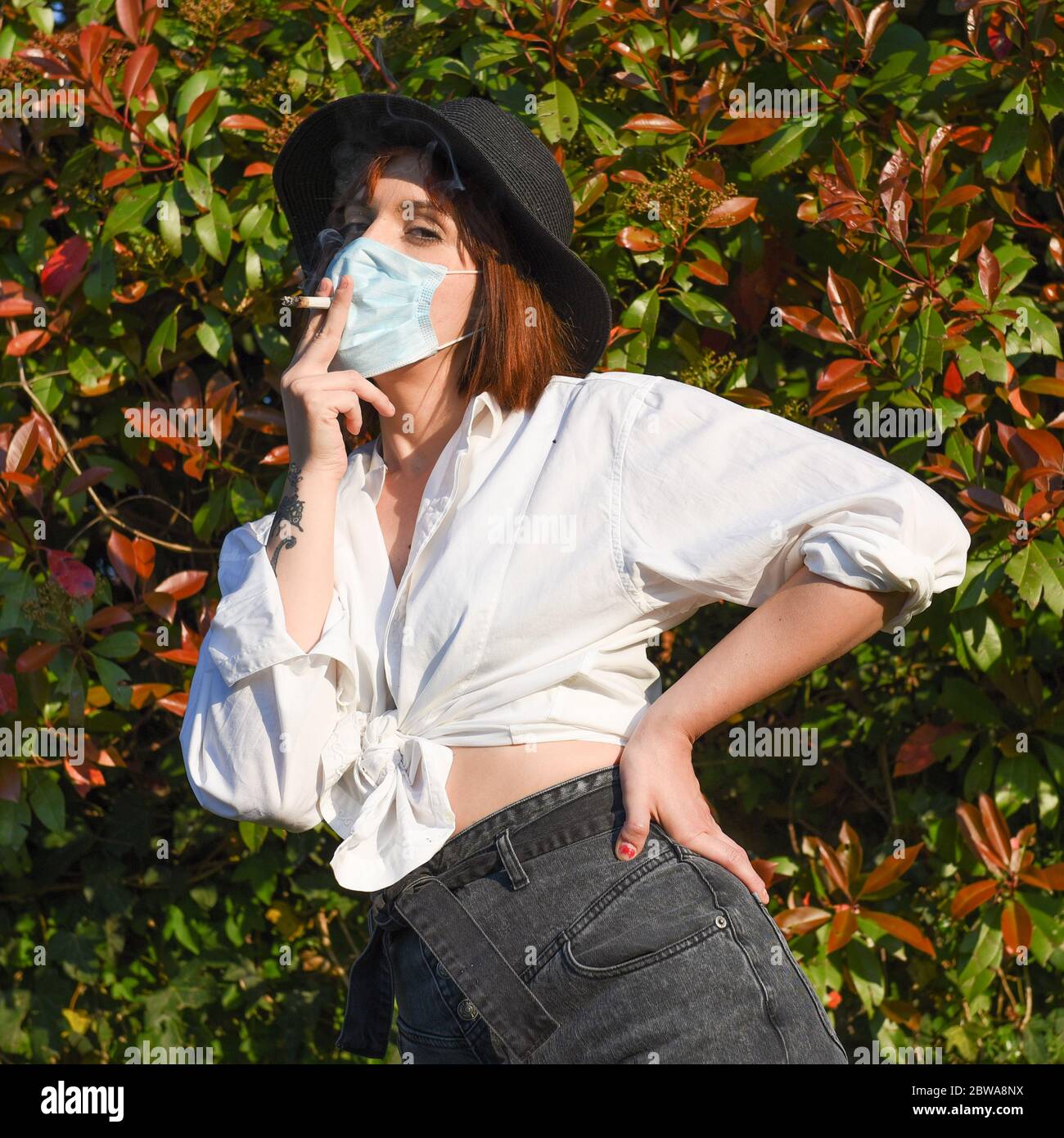 Girl with surgical face protection mask on the natural background smoking a cigarette. Stock Photo