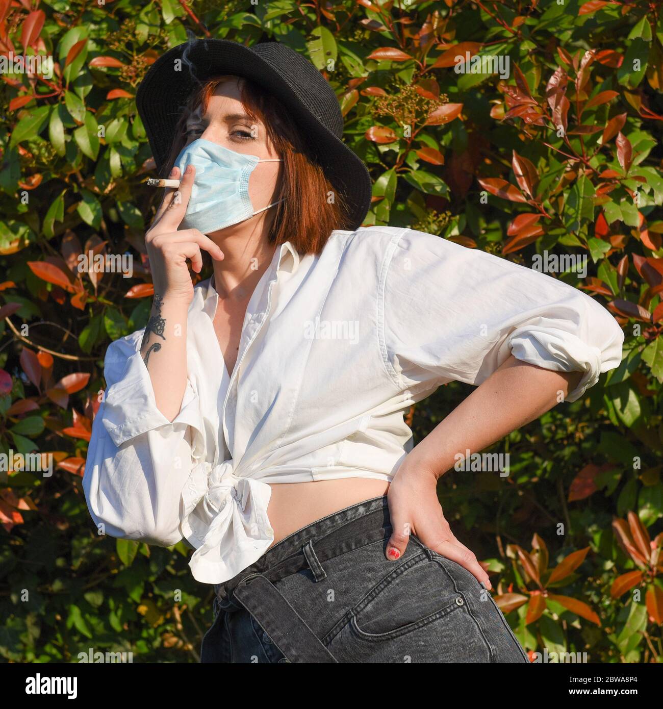 Girl with surgical face protection mask on the natural background smoking a cigarette. Stock Photo