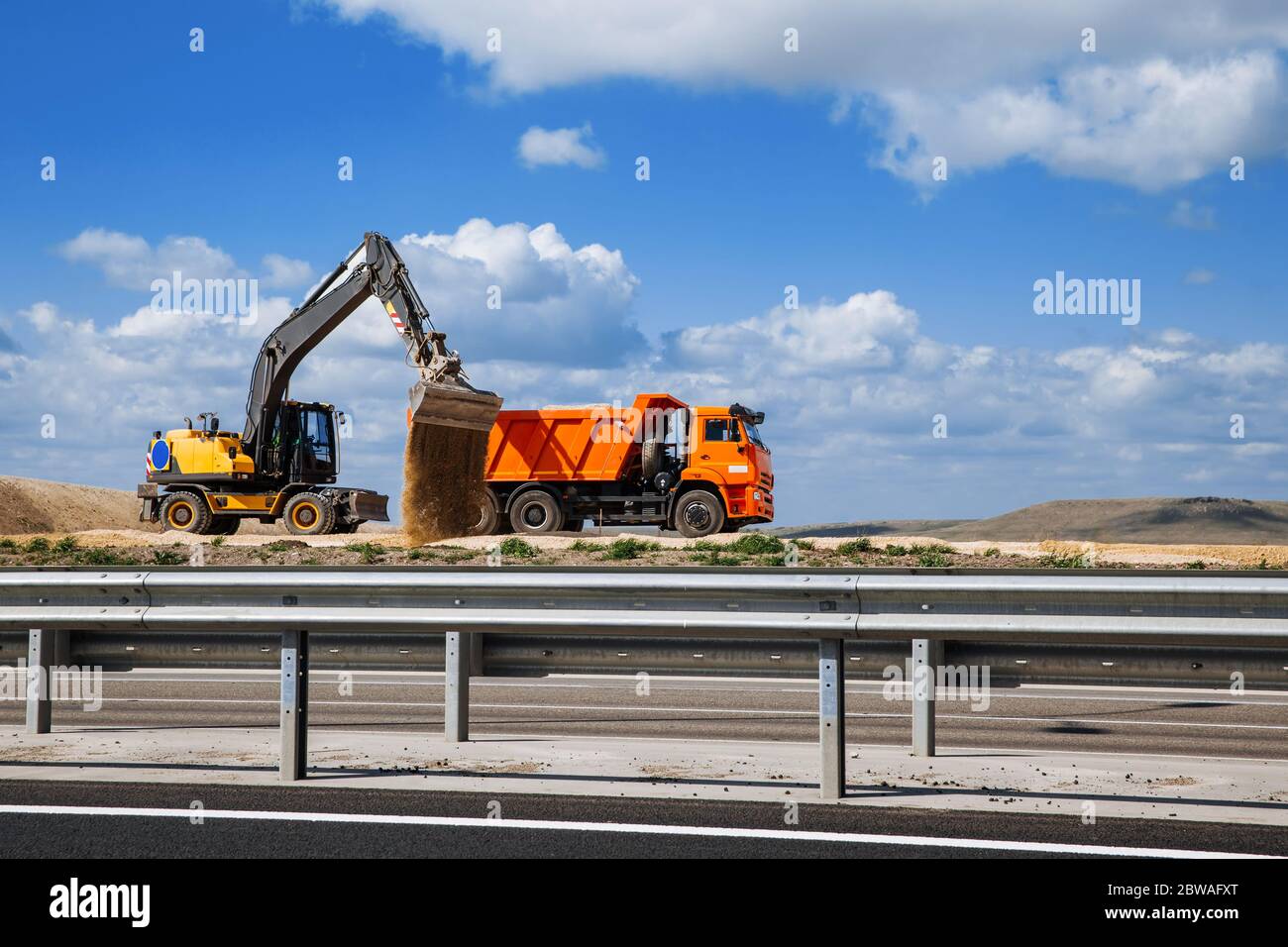 A yellow excavator pours sand from a large truck onto the road with a bucket. Road construction. Stock Photo