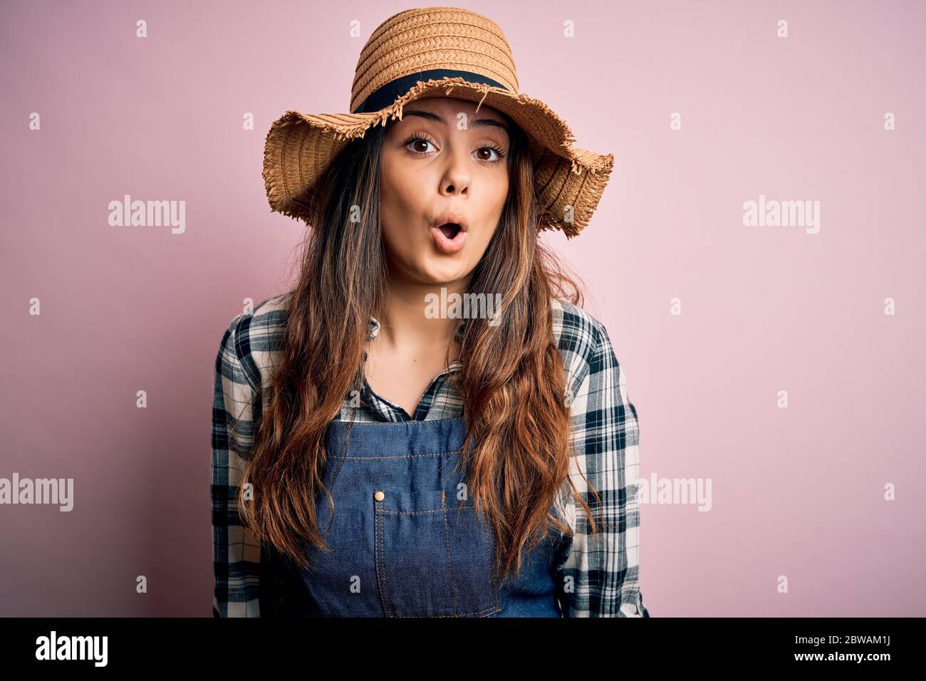 Young beautiful brunette farmer woman wearing apron and hat over pink ...