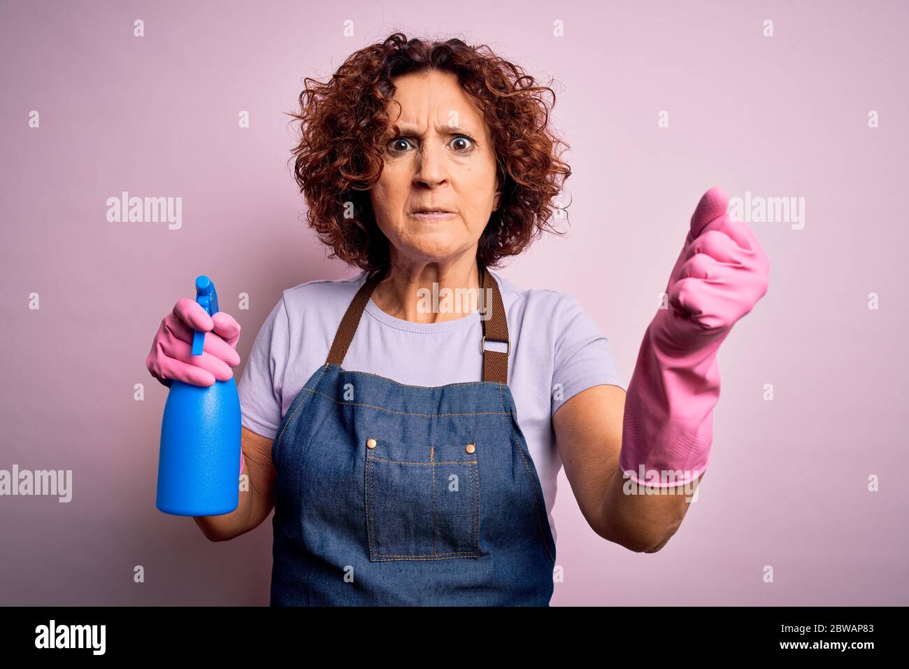 Middle age curly hair woman cleaning doing housework wearing apron and gloves using spayer annoyed and frustrated shouting with anger, crazy and yelli Stock Photo