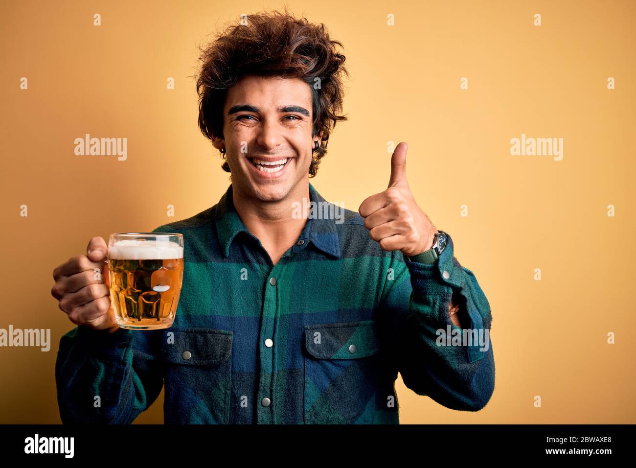 Young handsome man drinking jar of beer standing over isolated yellow ...