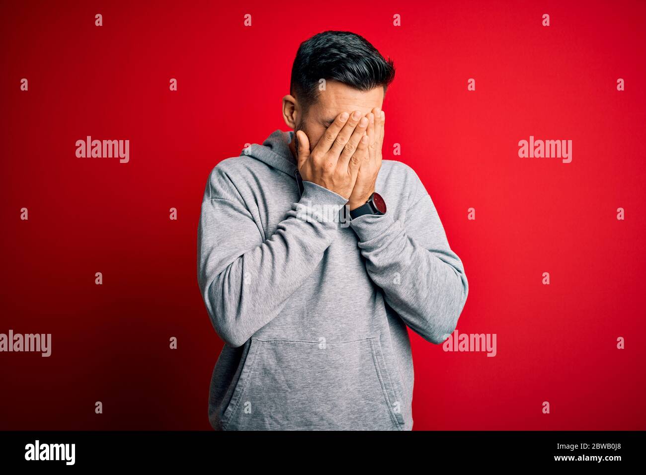 Young handsome sportsman wearing sweatshirt standing over isolated red background with sad expression covering face with hands while crying. Depressio Stock Photo