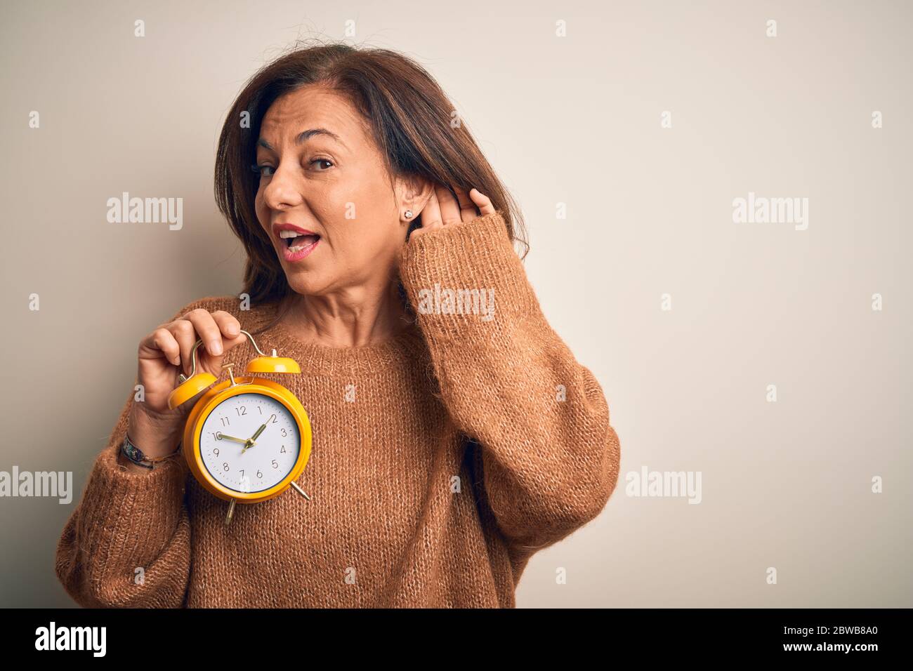 Middle age brunette woman holding clasic alarm clock over isolated background smiling with hand over ear listening an hearing to rumor or gossip. Deaf Stock Photo