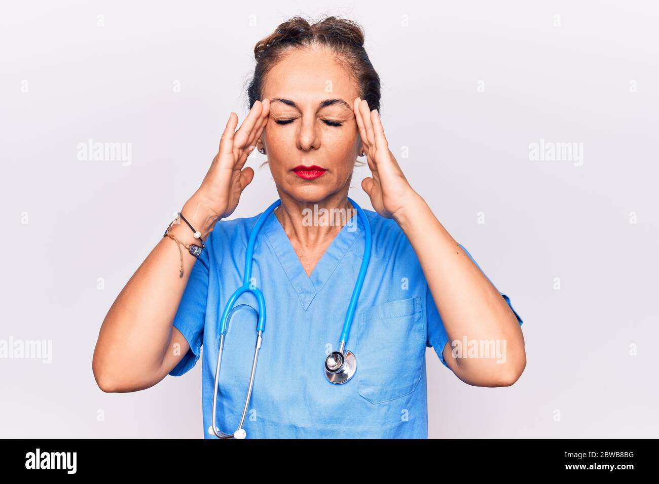 Middle age brunette nurse woman wearing uniform and stethoscope over white background with hand on head, headache because stress. Suffering migraine. Stock Photo