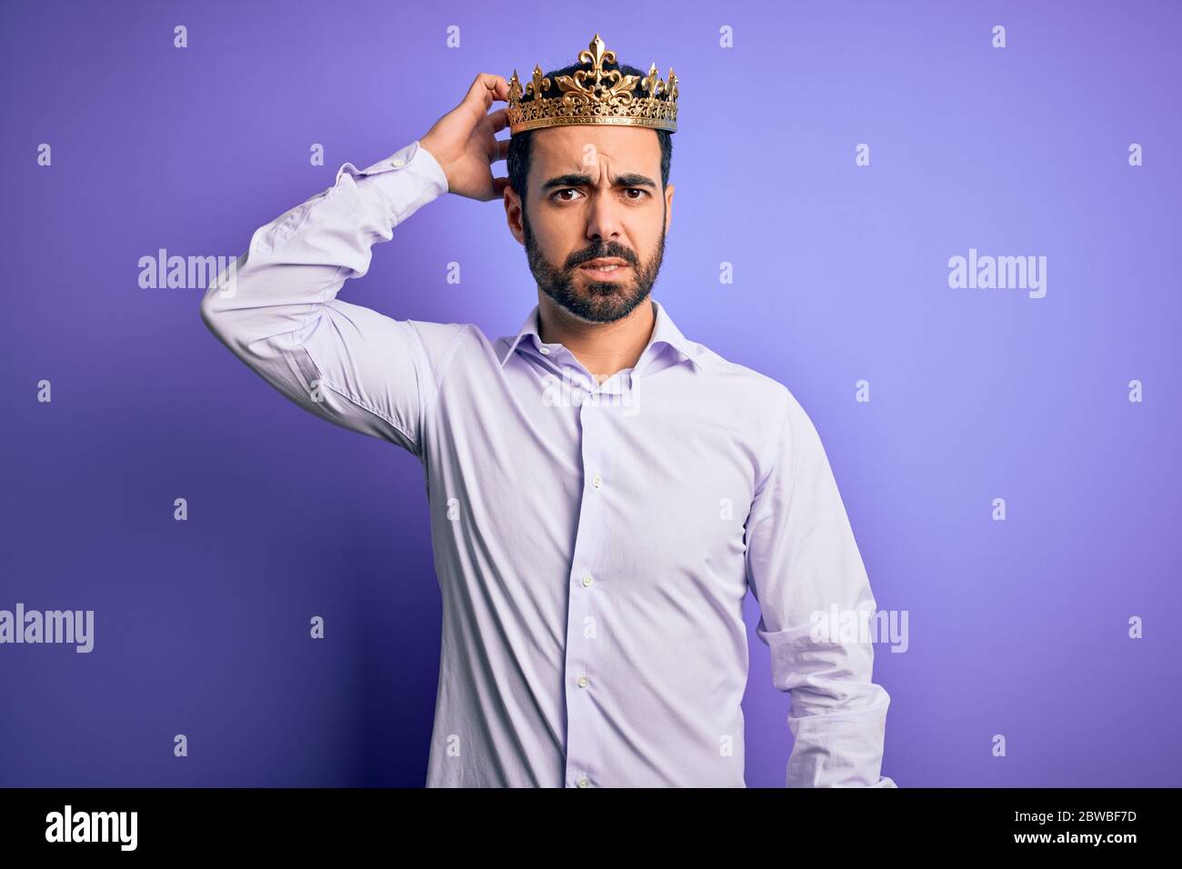 Young handsome man with beard wearing golden crown of king over purple ...