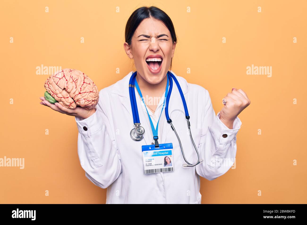 Young beautiful psychiatrist woman wearing stethoscope holding brain over yellow background screaming proud, celebrating victory and success very exci Stock Photo
